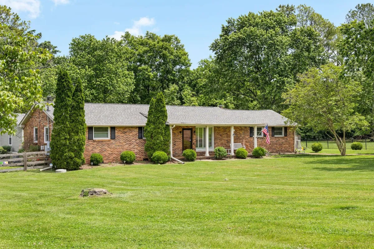 A wide view of the home's exterior showcases the brick facade, manicured lawn, and tall hedges, with an American flag waving gently. The expansive lawn and greenery set the scene for outdoor relaxation or picnics as the sun sets.