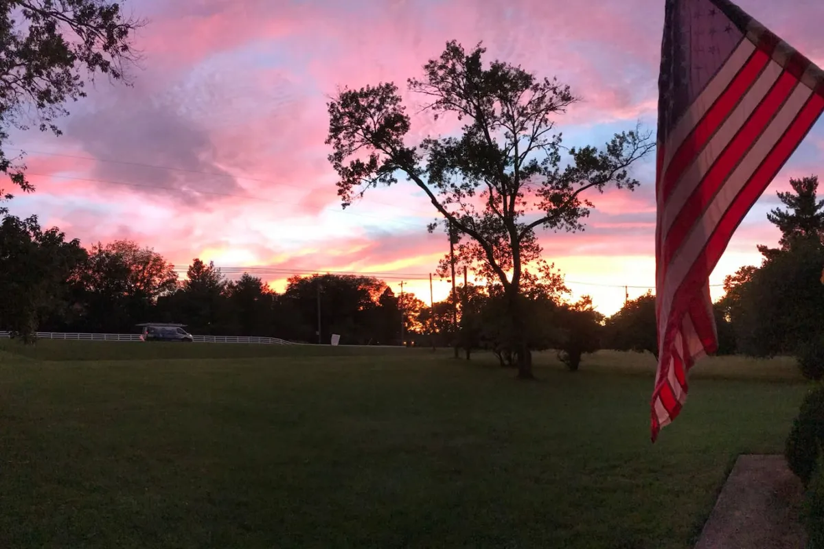 A breathtaking sunset fills the sky with warm hues, silhouetting trees and an American flag in the foreground. The tranquil scene captures the calm and beauty of the countryside as dusk settles in, inviting viewers to imagine quiet evenings outdoors.