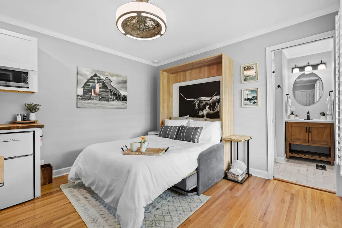 A relaxing and comfortable studio view showing a cozy bed with layered cushions and throws, artwork of a barn above the headboard, kitchenette amenities, and the entrance to the bathroom. Gentle daylight fills the room, emphasizing the versatility and warmth of the space.
