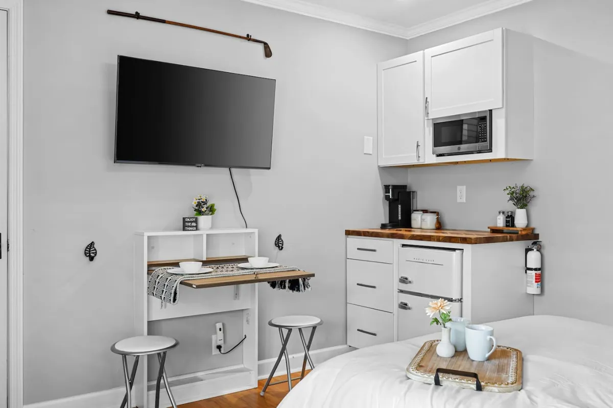 This image presents a compact kitchenette area with a wall-mounted TV, a small dining table with seating, and modern appliances. The setup is practical, maximizing use of space while remaining inviting and stylish. The table is positioned conveniently for enjoying meals, working, or relaxing, and the kitchenette includes smart storage solutions and sleek fixtures.