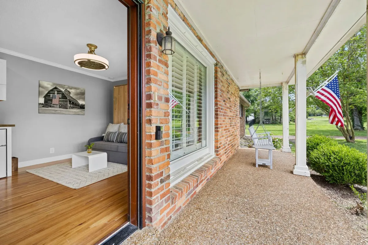 This image showcases the home's brick exterior with a welcoming front porch adorned by an American flag. Comfortable seating hints at a space for relaxing, while the lush yard and open skies suggest opportunities for enjoying both the sunrise and sunset from the porch.