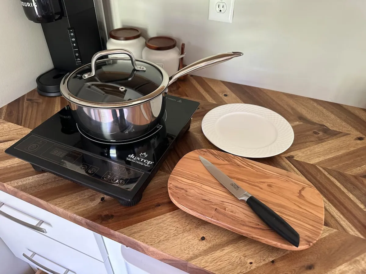 A focused view of a kitchenette workspace with an induction cooktop, wooden cutting board, knife, and white plate. The overall atmosphere is warm and functional, highlighting the essentials needed for preparing simple meals. Modern fixtures and neatly organized surfaces reinforce the sense of smart, well-planned amenities.