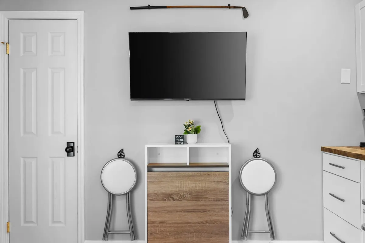 A modern corner of the studio featuring two round white chairs, a decorative plant, a wooden storage unit, and a TV mounted on the wall. The area is designed for relaxation, quiet contemplation, or socializing, with contemporary touches and soft, indirect lighting.