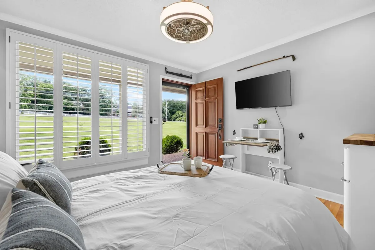 A serene studio space featuring a plush bed with white linens and decorative pillows, a small workspace with a chair in the corner, a television mounted on the wall, and an open door that frames a glimpse of the outdoors. The atmosphere is airy and calm, bathed in soft natural light, with the workspace and bed subtly separated by the room's layout.