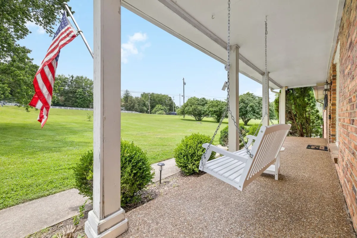 A peaceful view of the front porch featuring a swinging bench, American flag, and a sweeping, lush green lawn. The cozy bench invites relaxation, and the expansive lawn sets the stage for watching fireflies or sunsets in the evening.