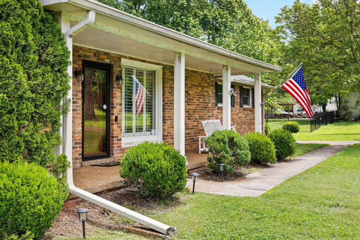 A welcoming front porch adorned with an American flag overlooks manicured bushes and a classic brick facade. The inviting outdoor space offers seating and is bathed in gentle evening light, conjuring ideas of quiet moments spent together as night falls.